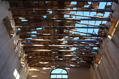Severely deteriorated roof structure with exposed wooden rafters and open sky visible through missing roof decking